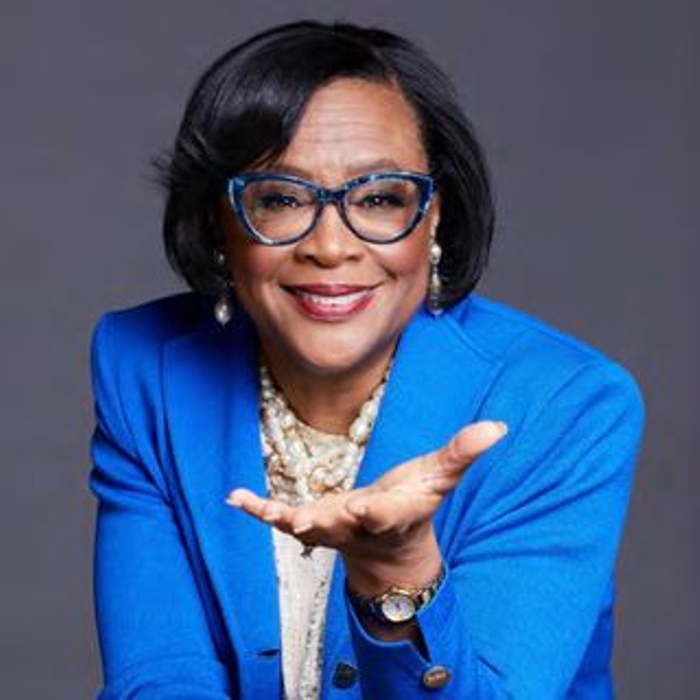 Cynt Marshall, extending hand. She is wearing glasses, a light blue suit jacket, and statement jewelry