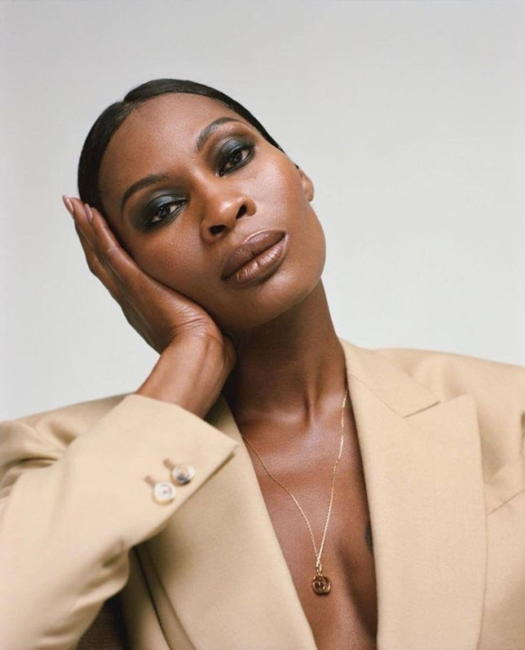 Dominique Jackson posed with one hand on face, wearing a tan blazer and pendant necklace