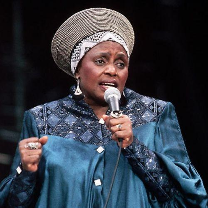 Miriam Makeba singing. She is wearing a headdress and shirt with geometric patterns.