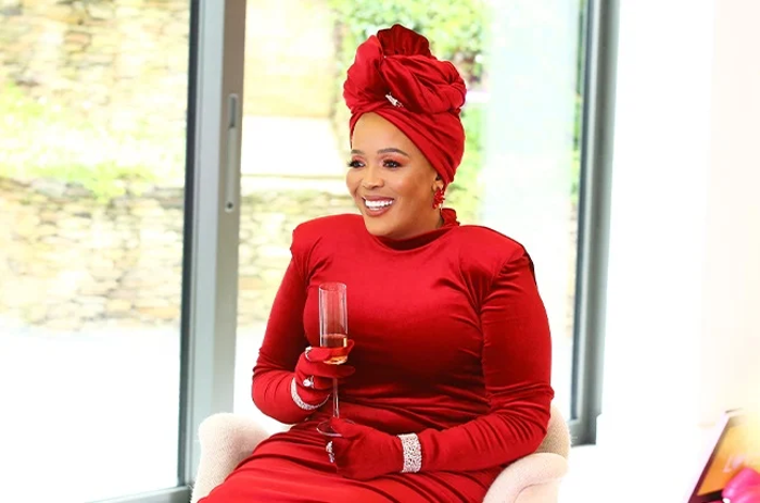 Tumi Morake seated and holding a champagne flute. She is wewaring a red headwrap, matching dress, and diamond bracelets and rings