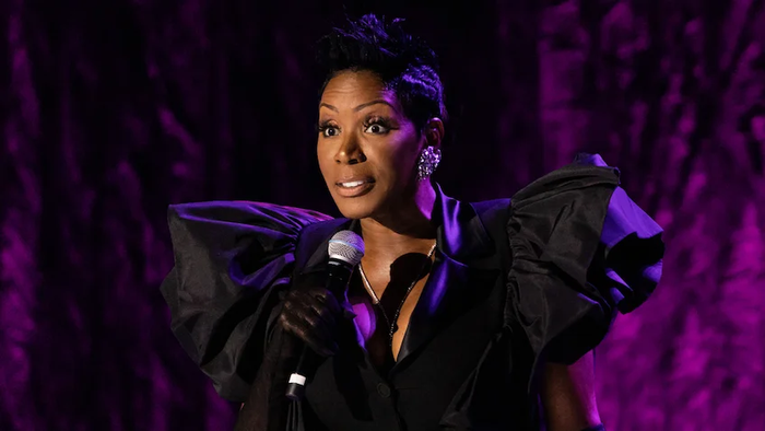 Sommore performing with microphone, she is wearing a black dress with puffy sleeves and sparkling statement earrings