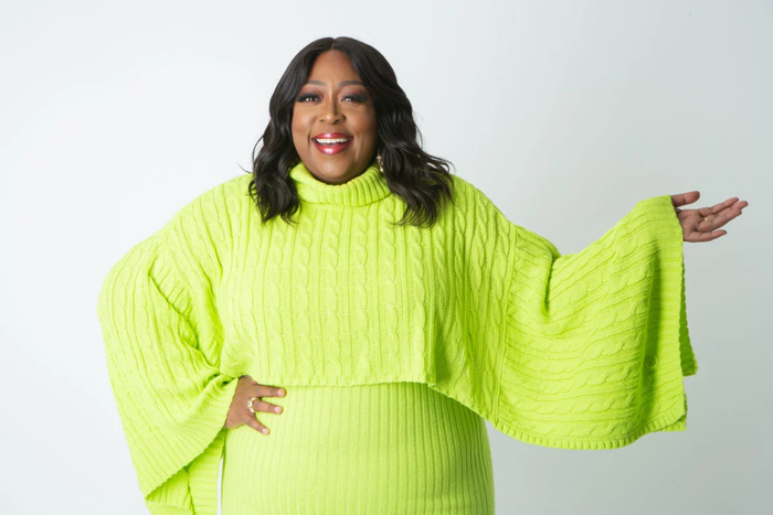 Loni Love, smiling posed with arm outstretched and other arm on her hip.