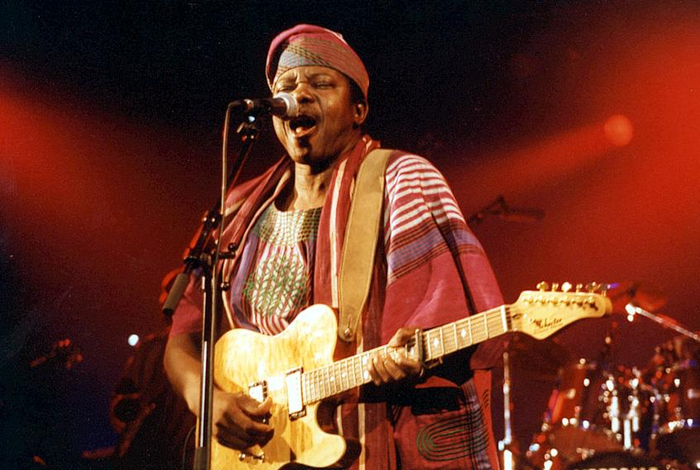 King Sunny Ade in traditional clothing and hat, playing guitar on stage