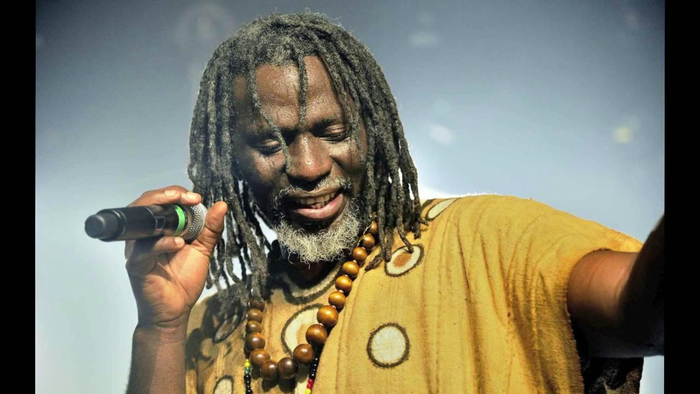 Tiken Jah Fakoly, in locs and beard, and wooden beads around his neck, and yellow patterned kaftan performing on stage