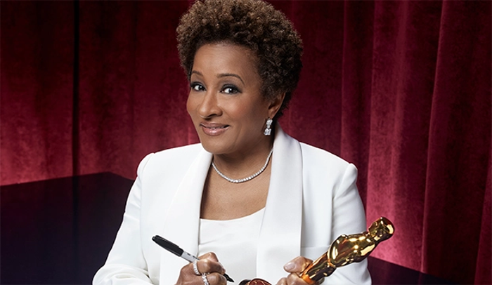 Wanda Sykes in a white suit jacket and top signing an Oscar with a confident smile