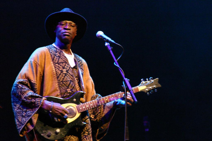 Ali Farka Toure, in black wide brimmed hat, traditional patterened robe, and glasses, playing guitar on stage