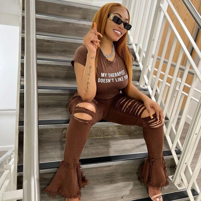 Lisa Mercedez sitting on stairs in brown shirt that says "My Therapist Doesn't Like You" and brown ripped bell bottom jeans. She is wearing sunglasses and a silver chunky chain.