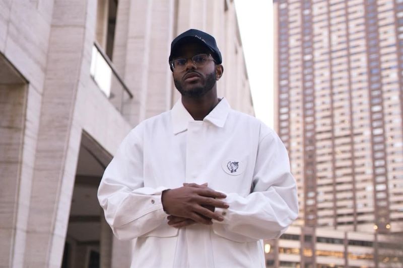 Chef Kwame Onwuachi in baseball hat and chef's coat, hands folded in front, in the city