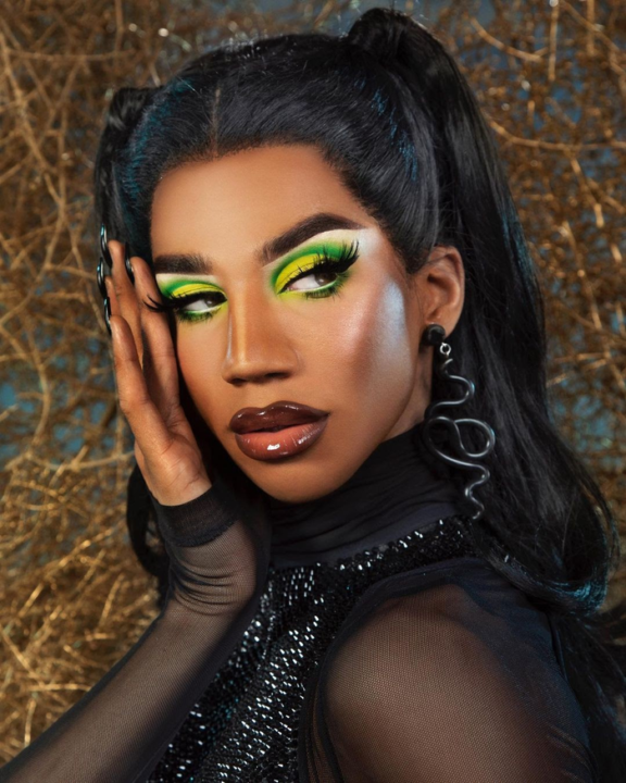 Naomi Smalls posing with one hand on face, she is wearing a mesh shirt with sparkling beads and statement earrings, her hair is in pigtails 