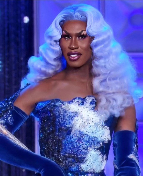 Shea Coulee in white wavy wig, dramatic makeup, and a royal blue dress covered in sequins and intricate patterns with matching gloves