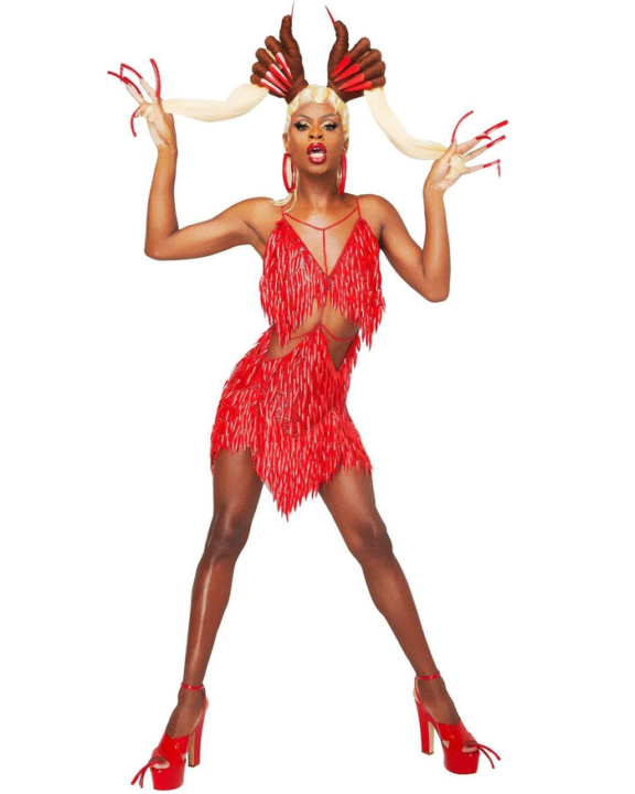 Symone is wearing a fringe cutout dress in red with super long nails and hair that is also fashioned with an accessory shaped like hand with long nails. He is wearing red platform heels.