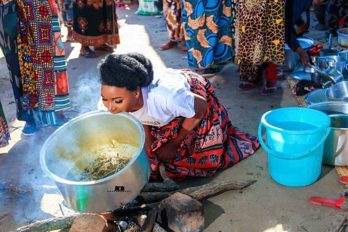 Bride lifting large pot over fire with her teeth,  crouched down. Her hands are in front of her.