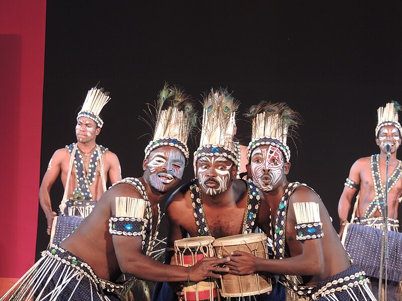 Dancers in ornate headdresses with peacock feathers and cowry shells, and cowry shell decorated arm bands and straps and skirts, they are wearing facepaint with geometric designs, and singing and playing drums.