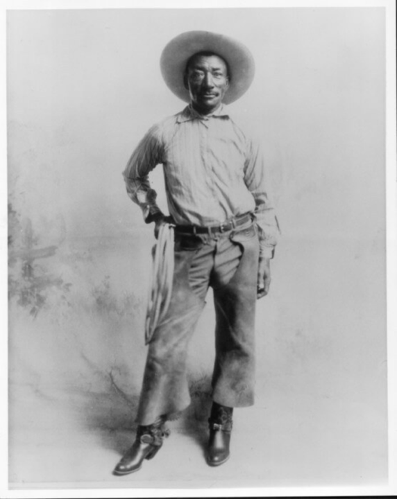 Black and white photo of Bill Pickett, with right hand on hip holding lasso, chaps, and cowboy hat and boots with spurs.