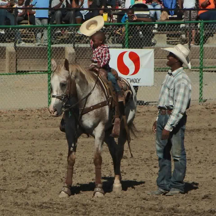 a little Black boy riding on a pony and waving cowboy hat at rodeo. The horse trainer, also an African American cowboy, stands close by.