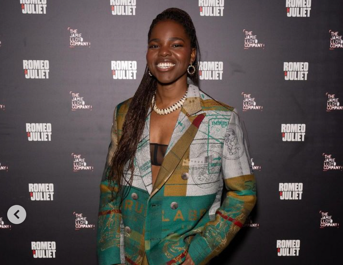 Francesca Amewudah-Rivers at an event promoting Romeo & Juliet, she is wearing gold hoop earrings, a tailored jacket with colorful patterns, and a sheer tube top underneath, and shell necklace.