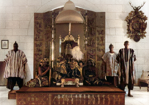 An Oyo king sitting on elbaorate throne with guards and a female attendant also in traditional attire. Sepia photo.