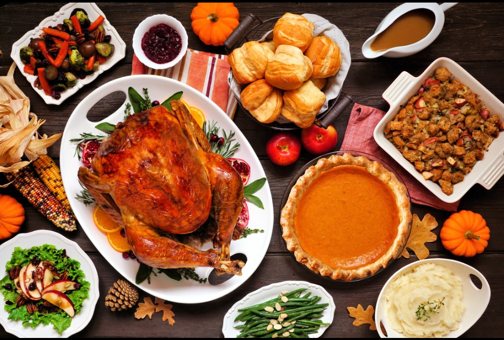 A full spread of Thanksgiving dinner including roasted turkey, dinner rolls, stuffing, green beans, mashed potatoes, roasted vegetables, and pumpkin pie.