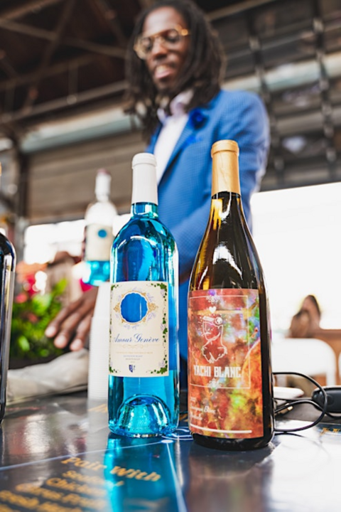 Black man in suit and locs reaching for a bottle of wine