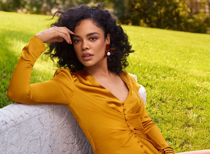 Tessa Thompson seated at a field wearing pearl earrings and a satin yellow button down dress