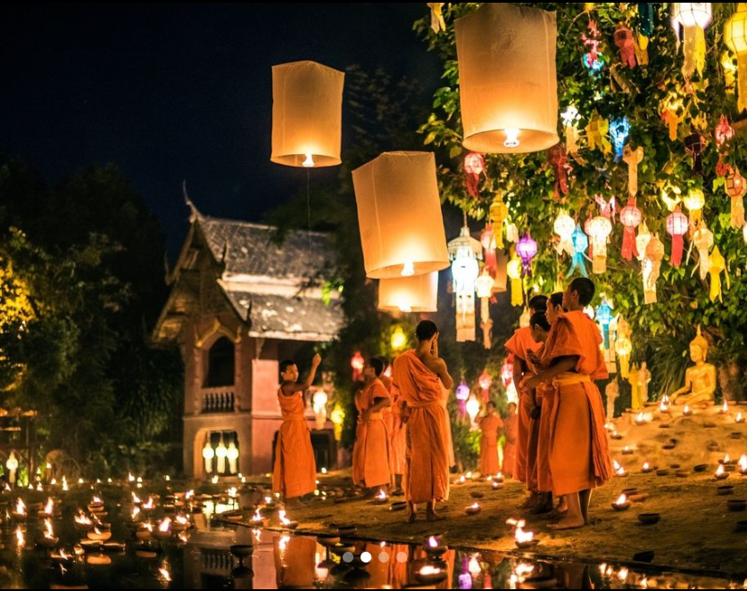 Young Thai monks in the night, under a tree decorated with colorful paper lanterns. They are sending a flying paper lantern into the sky, and the river bank where they are is also full of candlelight on the ground and on the water.