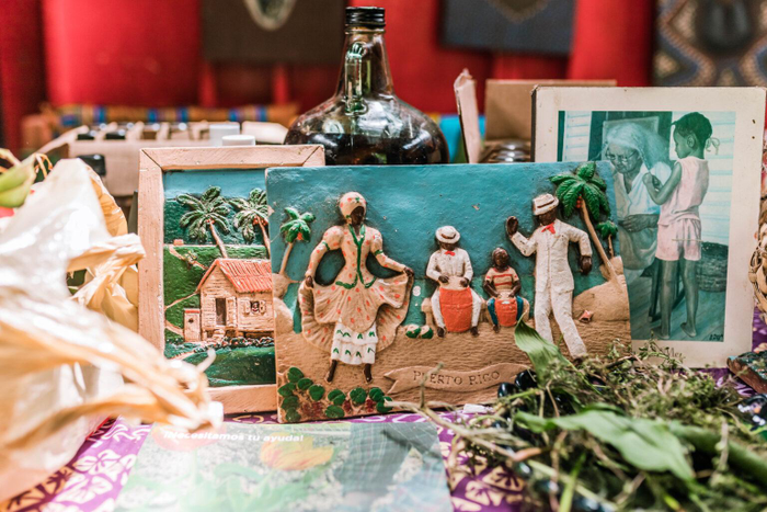 Framed photograph of Peurto Rican family, a relief of dancers, and a relief of a rustic house, arranged with a bottle and herbs set on a table