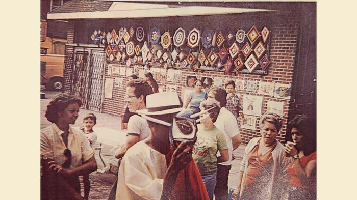 A faded color photograph of Puerto Rican immigrants in Pennsylvania decades ago. They are outside a building decorated with woven artwork.