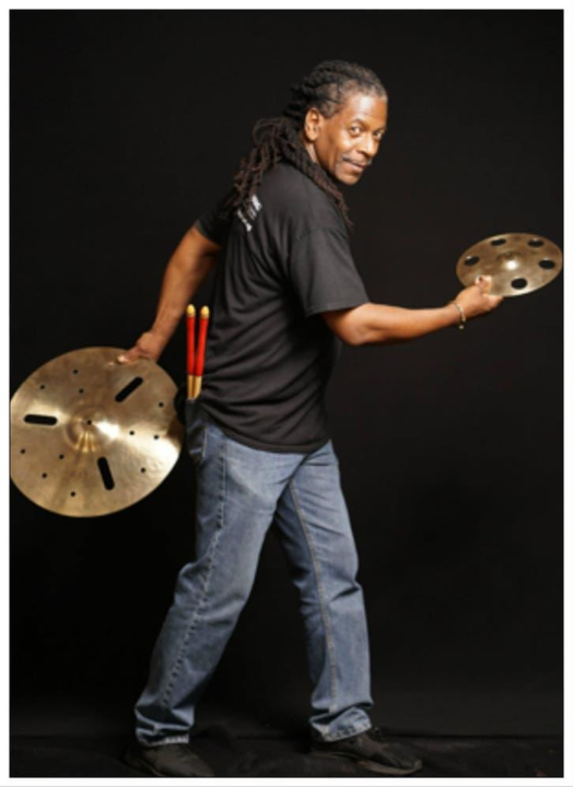 Man in dreadlocs posing with cymbals and percussion sticks in his back pocket.