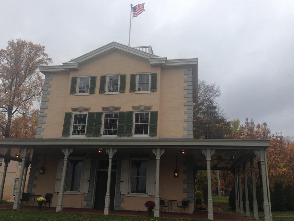 The Belmont Mansion, a colonial style building with a porch supported by multiple columns. The American flag stands at the top of the building.