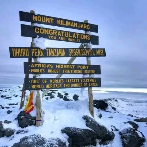 Wooden sign at the snow-covered summit of Mount Kilimanjaro reading “Mount Kilimanjaro – Congratulations, you are now at Uhuru Peak, Tanzania, 5895m / 19341ft AMSL,” with additional text describing it as Africa’s highest point, the world’s highest free-standing mountain, one of the largest volcanoes, and a World Heritage site.