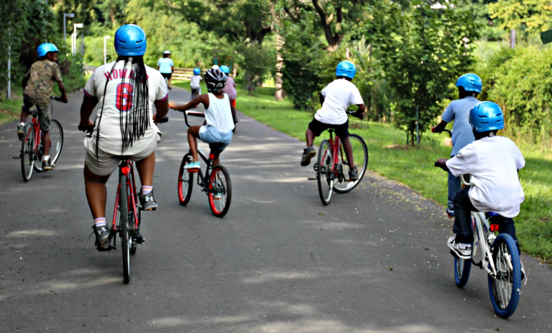 group of children and adult wearing helmets on bikes in a  path surrounded by greenery