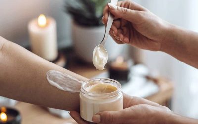 pair of hands holding a jar of shea butter and a spoon and applying it to someone's arm