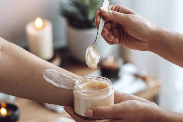 pair of hands holding a jar of shea butter and a spoon and applying it to someone's arm