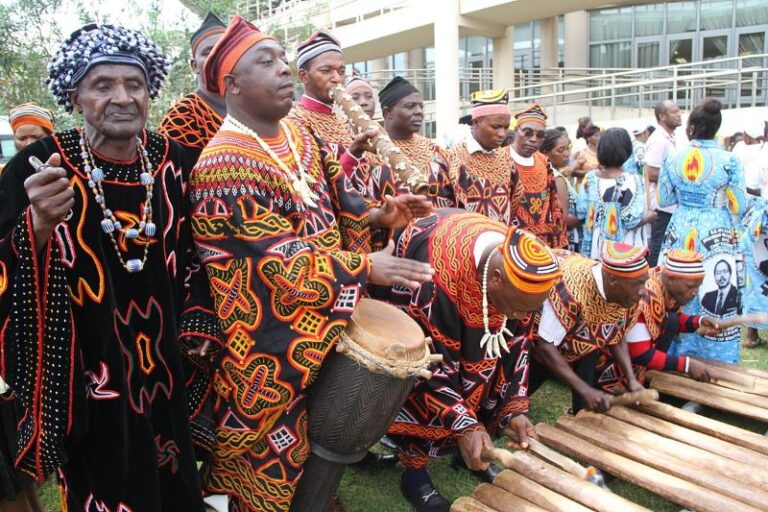 Cameroonian musicians in colorful traditional attire playing drums, wooden percussion instruments, and a wind instrument.