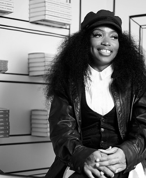 Jeannine Cook seated inside bookshop, smiling. She is wearing a hat, and leather jacket over a collared shirt and vest