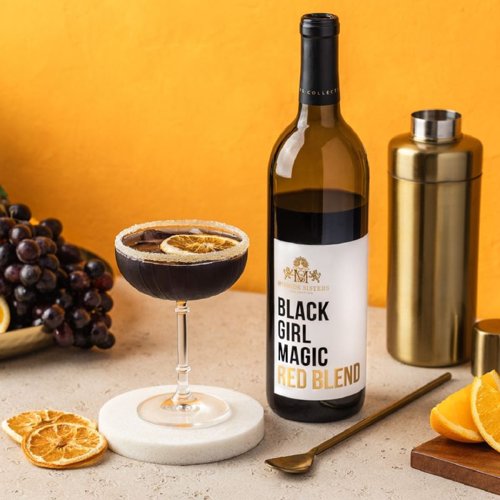 A bottle of Black Girl Magic Red Blend wine, featured with a wine cocktail in martini glass rimmed with sugar and a dried orange floating on top