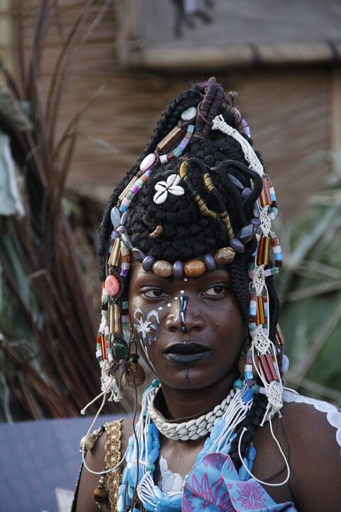 Woman wearing detailed beaded headdress with shells and two-tiered shell necklace, and face paint. Her hair is in braids on top of her head.