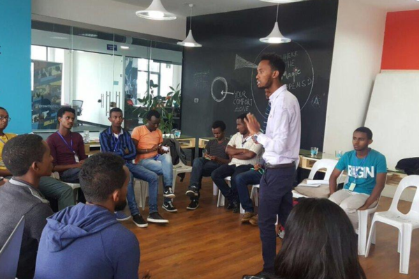 A meeting at a startup company in Ethiopia with a giant chalkboard wall in background and one presenter. Other employees are seated in a circle around him.