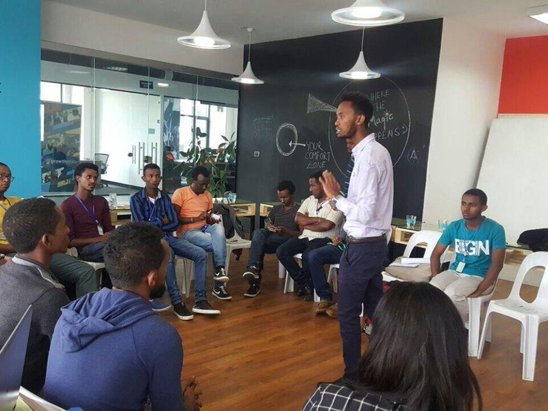 A meeting at a startup company in Ethiopia with a giant chalkboard wall in background and one presenter. Other employees are seated in a circle around him.