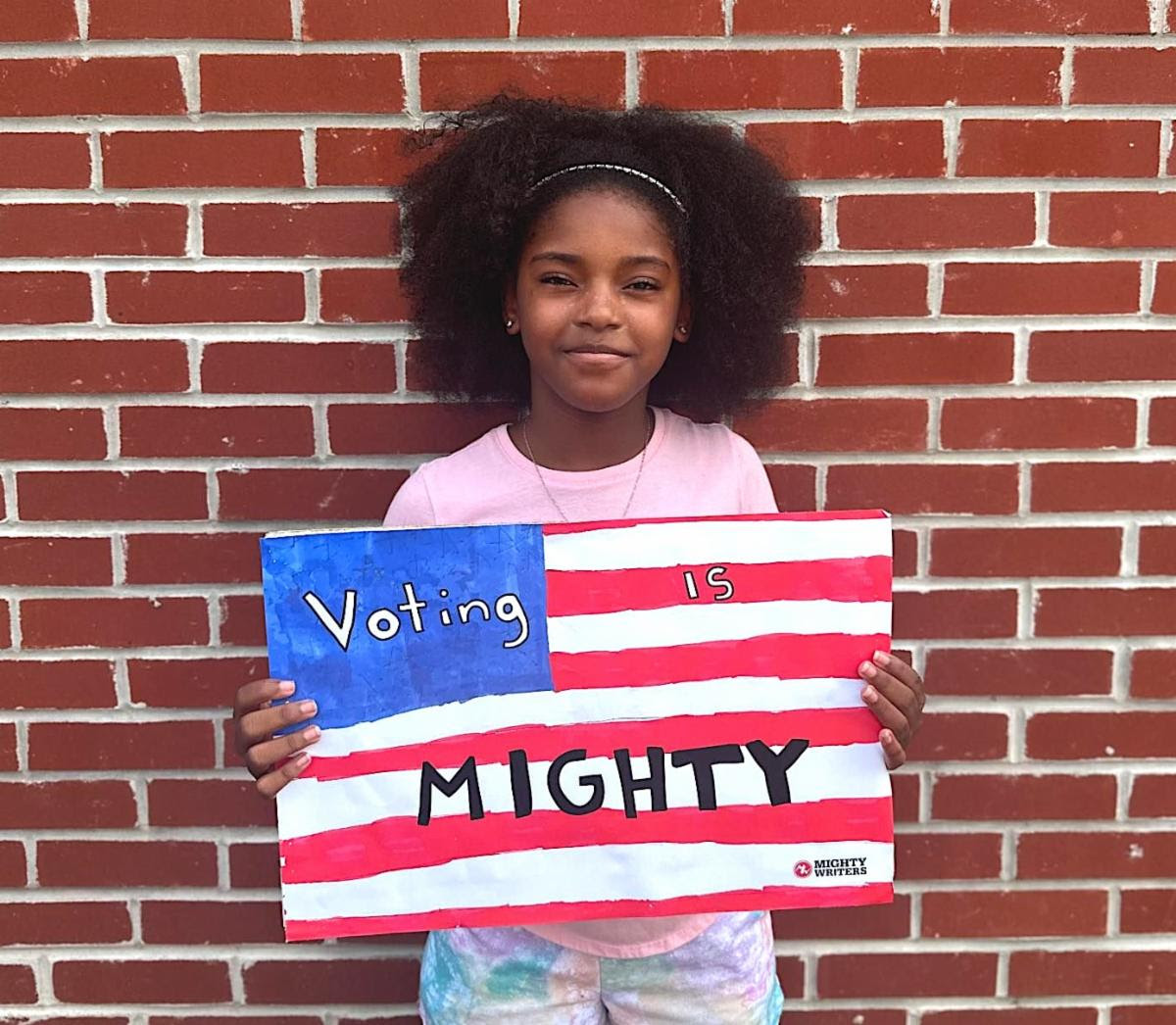 Young African American girl holding a handmade sign that looks like the American flag that says "Vote Mighty"