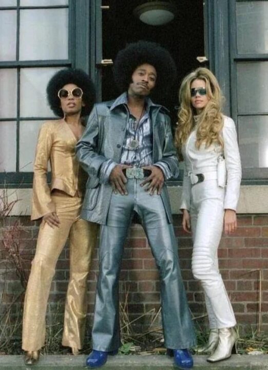 Eddie Griffin in 1970s style afro and jacket and bellbottom pants, with necklace and embellished belt. The tow ladies next to him are similarly dressed in 1970s style suits and heels.