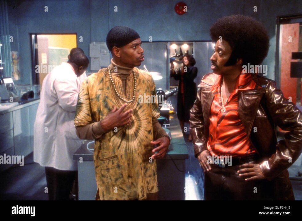 Dave Chapelle in durag, bead necklace, tie-die kaftan and turtleneck, talking to Eddie Griffin in afro, 1970s jacket and shirt and gold chain.