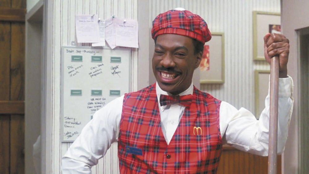 Eddie Murphy in plaid cap and matching vest and tie and white collared shirt, holding a mop.