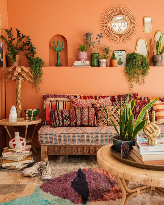 Maximalist house decoration with woven rattan table, potted plants, colorful throw cushions on couch with carved wooden frame, piled magazines, lots of interior decorations, and colorful rug