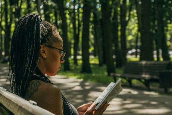 Black woman with locs in ponytail reading a book outdoors on a park bench surrounded by trees