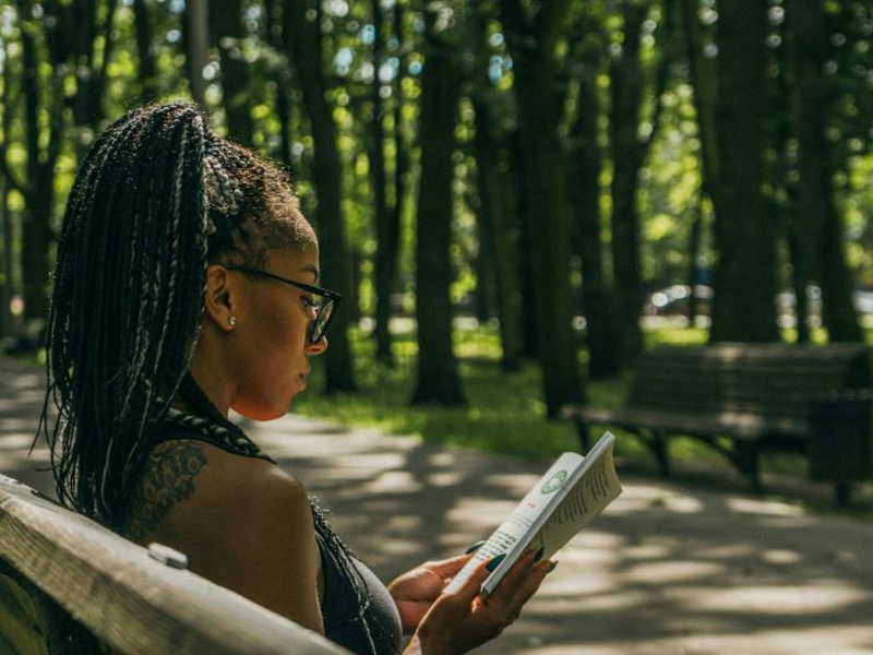 Black woman with locs in ponytail reading a book outdoors on a park bench surrounded by trees