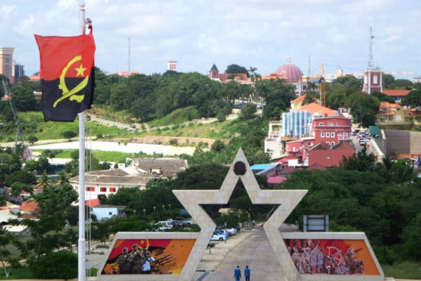 A stone arch shaped like a star, with colorful orange and red buildings with green hills and trees in the background. The Angola flag on a pole is in the foreground.