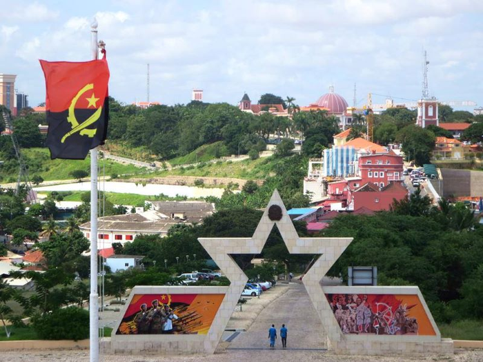 A stone arch shaped like a star, with colorful orange and red buildings with green hills and trees in the background. The Angola flag on a pole is in the foreground.