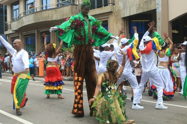 Performers at the Festival Kreol. In the middle is a stilts performer and in front is a dancer dressed as a fairy, and surrounding them are other dancers in matching colorful outfits.