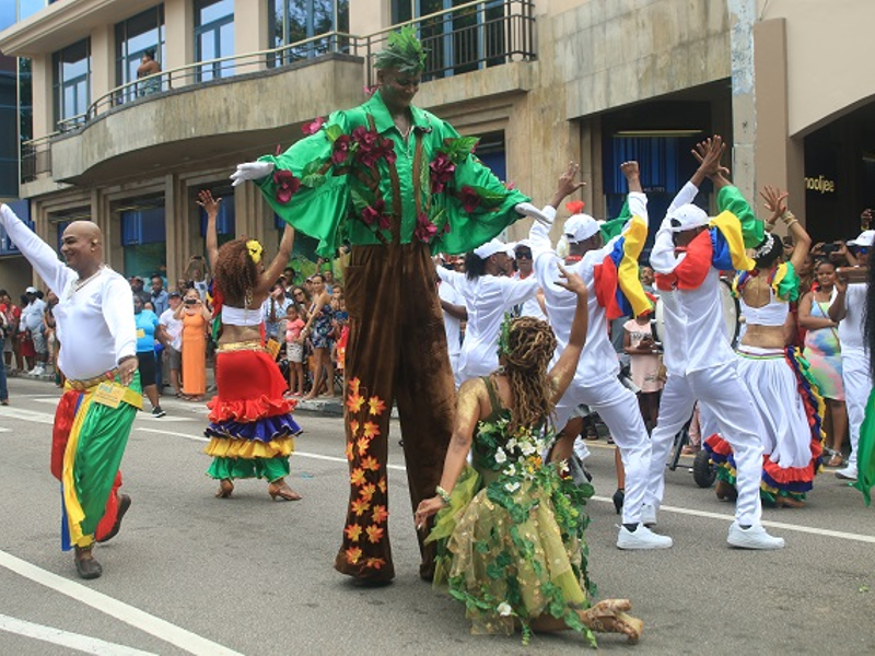 Performers at the Festival Kreol. In the middle is a stilts performer and in front is a dancer dressed as a fairy, and surrounding them are other dancers in matching colorful outfits.